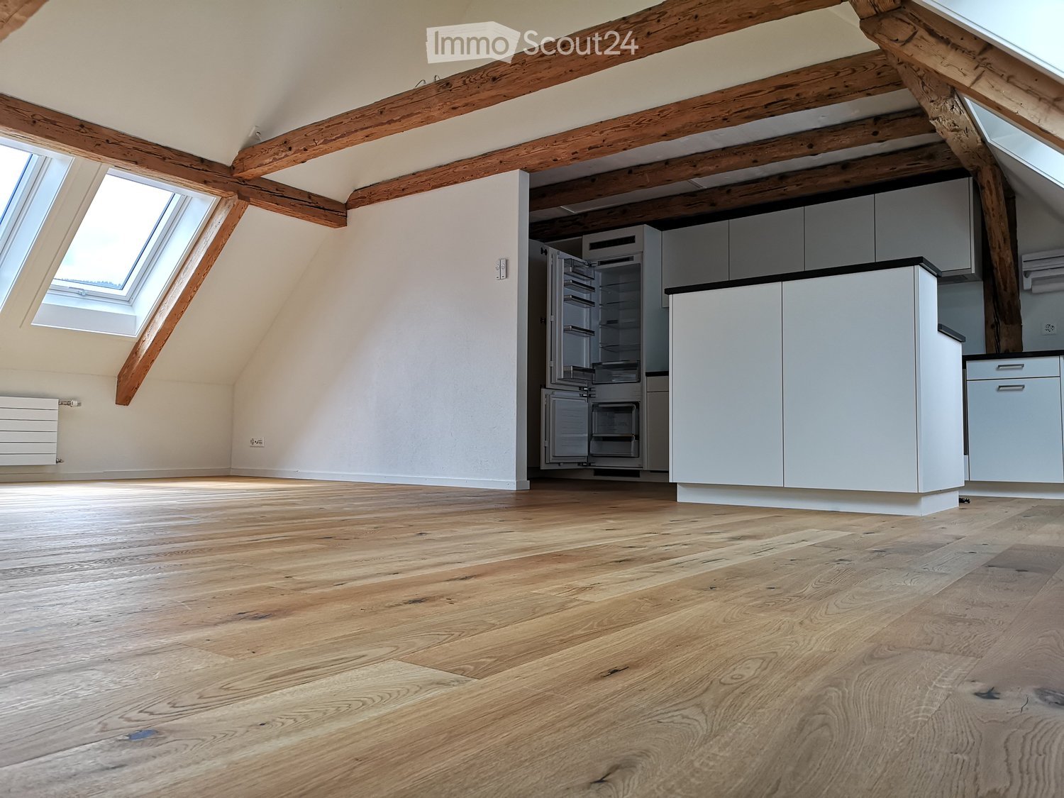Bright, airy kitchen with exposed wooden beams, white cabinets, and a modern refrigerator. The space features light wooden flooring and large skylight windows.
