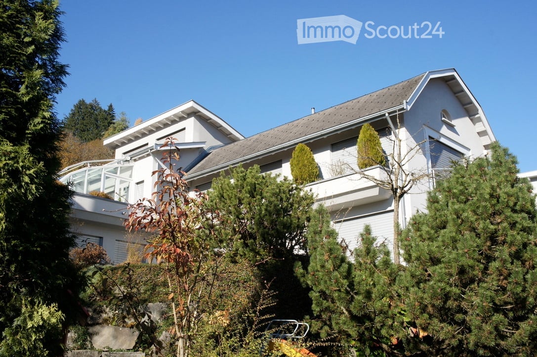 Two-story white house with a raised ground floor, surrounded by trees and shrubs.