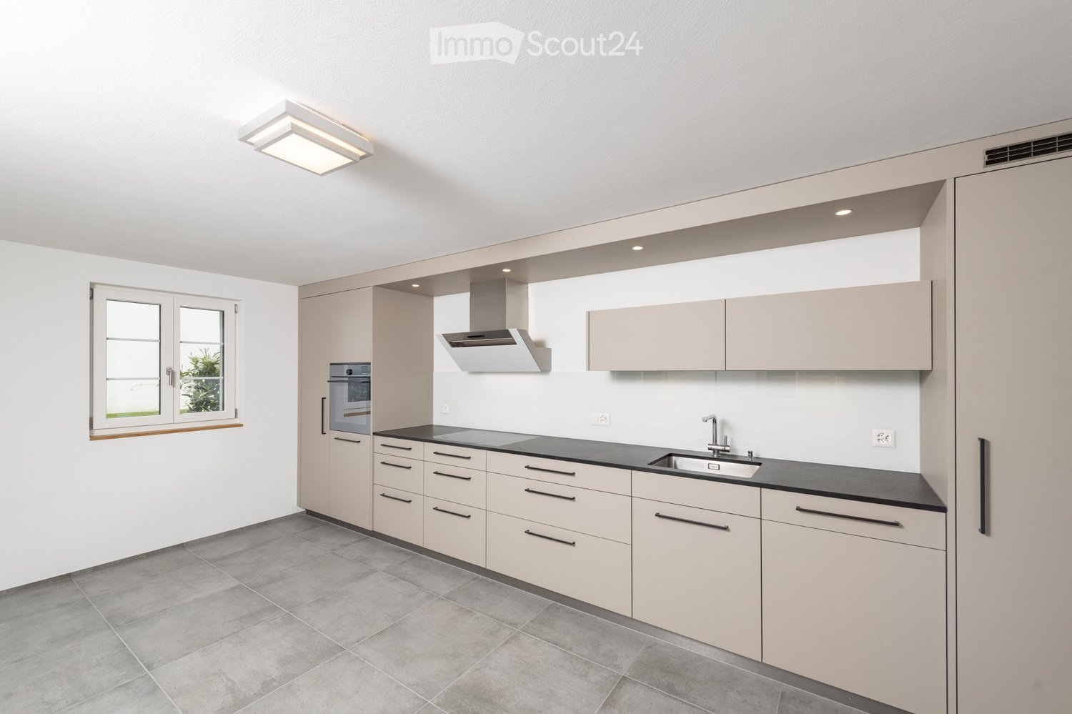 Modern kitchen with light-colored cabinets, black countertops, a sink with a faucet, an oven, and a range hood. The space is well-lit with recessed lighting and features a window with a view of greenery.
