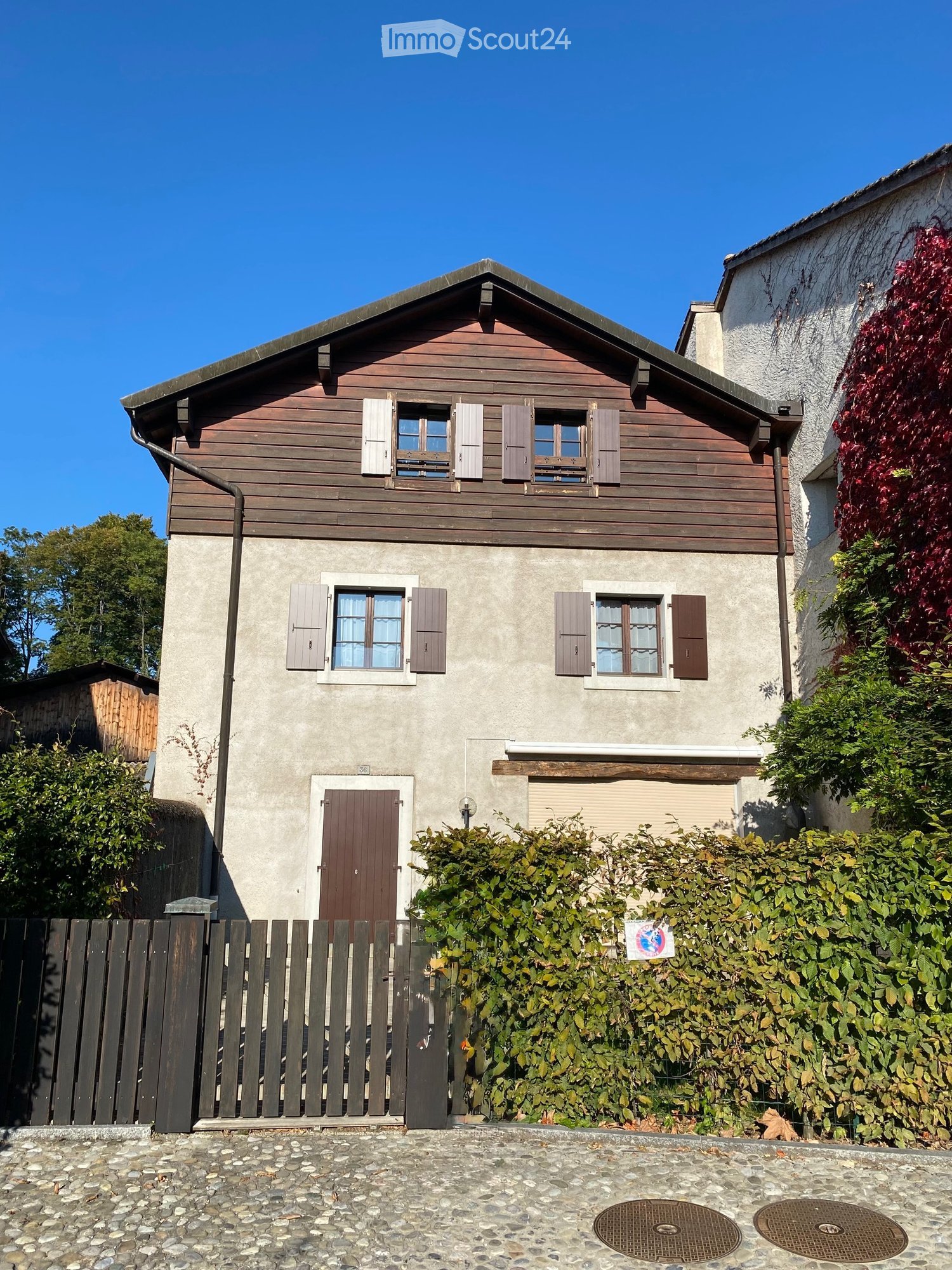 Two-story house with wooden siding on the upper level and concrete on the lower level, featuring multiple windows with shutters and a central front door.
