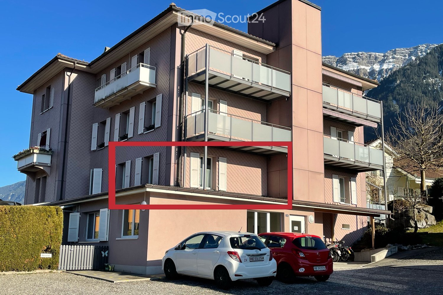 A multi-story residential building with balconies, white shutters, and a modern design, set against a backdrop of snow-capped mountains.