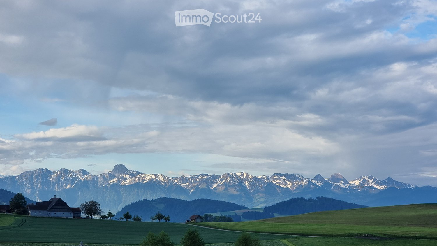 A serene rural landscape with green fields, scattered trees, a small house, and distant snow-capped mountains under a partly cloudy sky.