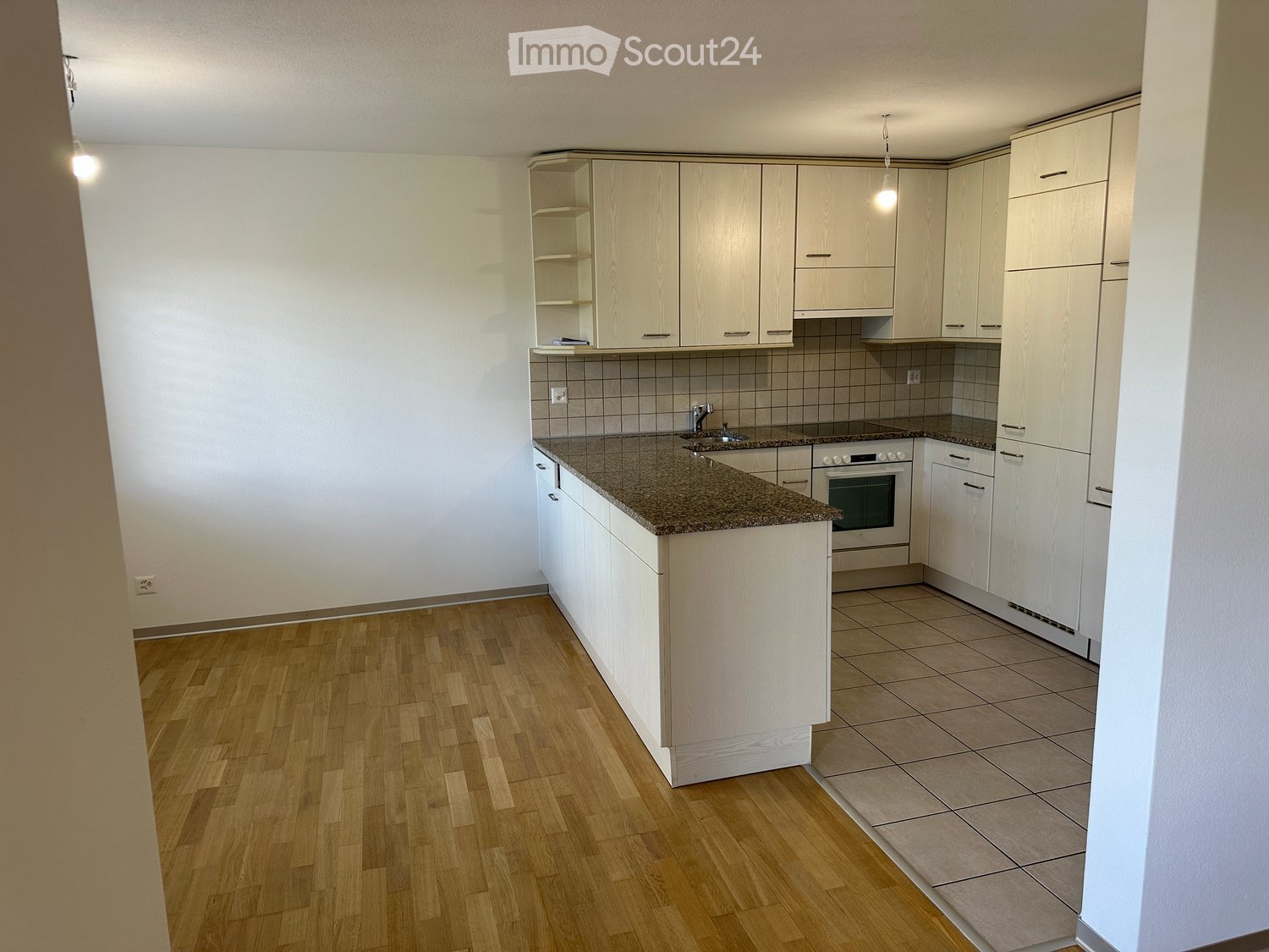 Modern kitchen with light wood cabinets, granite countertop, and built-in oven. Wooden flooring in the main area and tiled flooring near the sink.