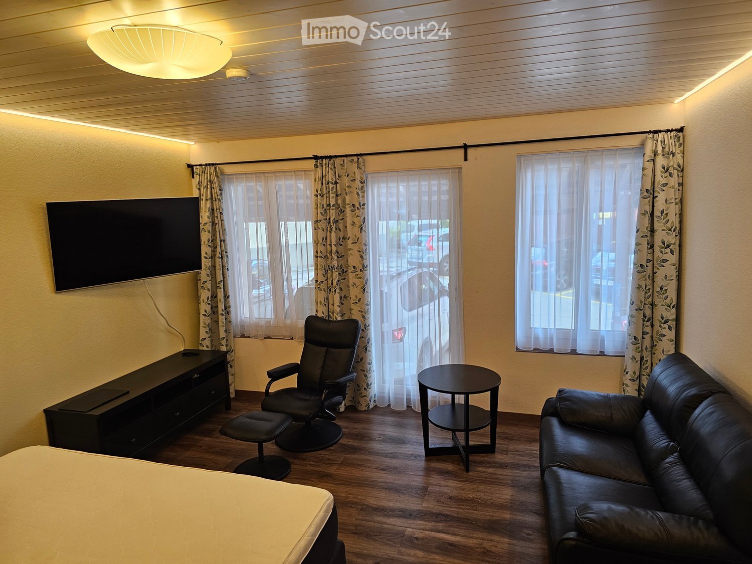 Living room with bed, couch, TV, desk, chair, side table, hardwood floor, yellow ceiling light
