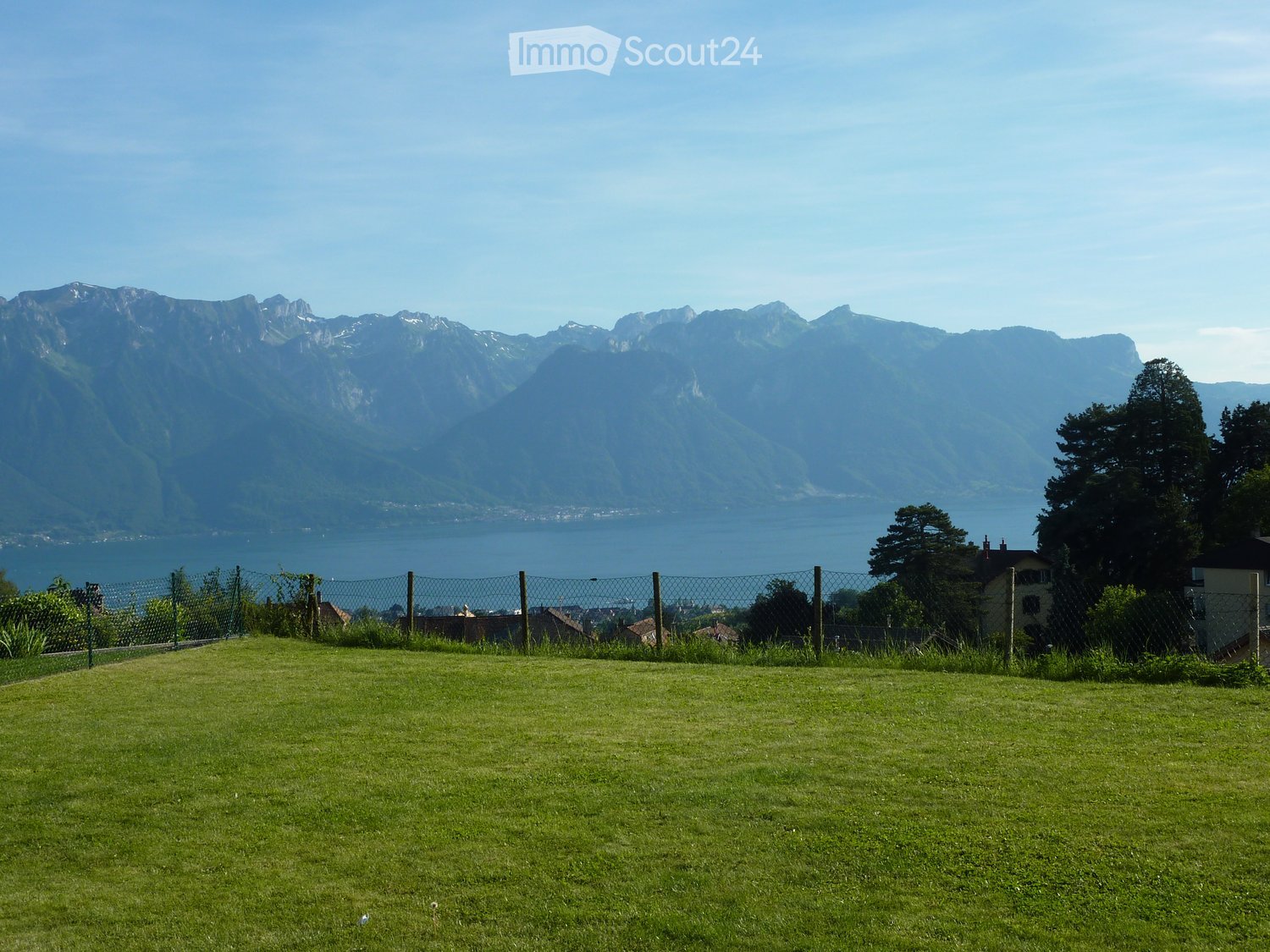 A grassy lawn with a metal fence, surrounded by trees and houses, with a scenic view of a lake and mountains in the background.