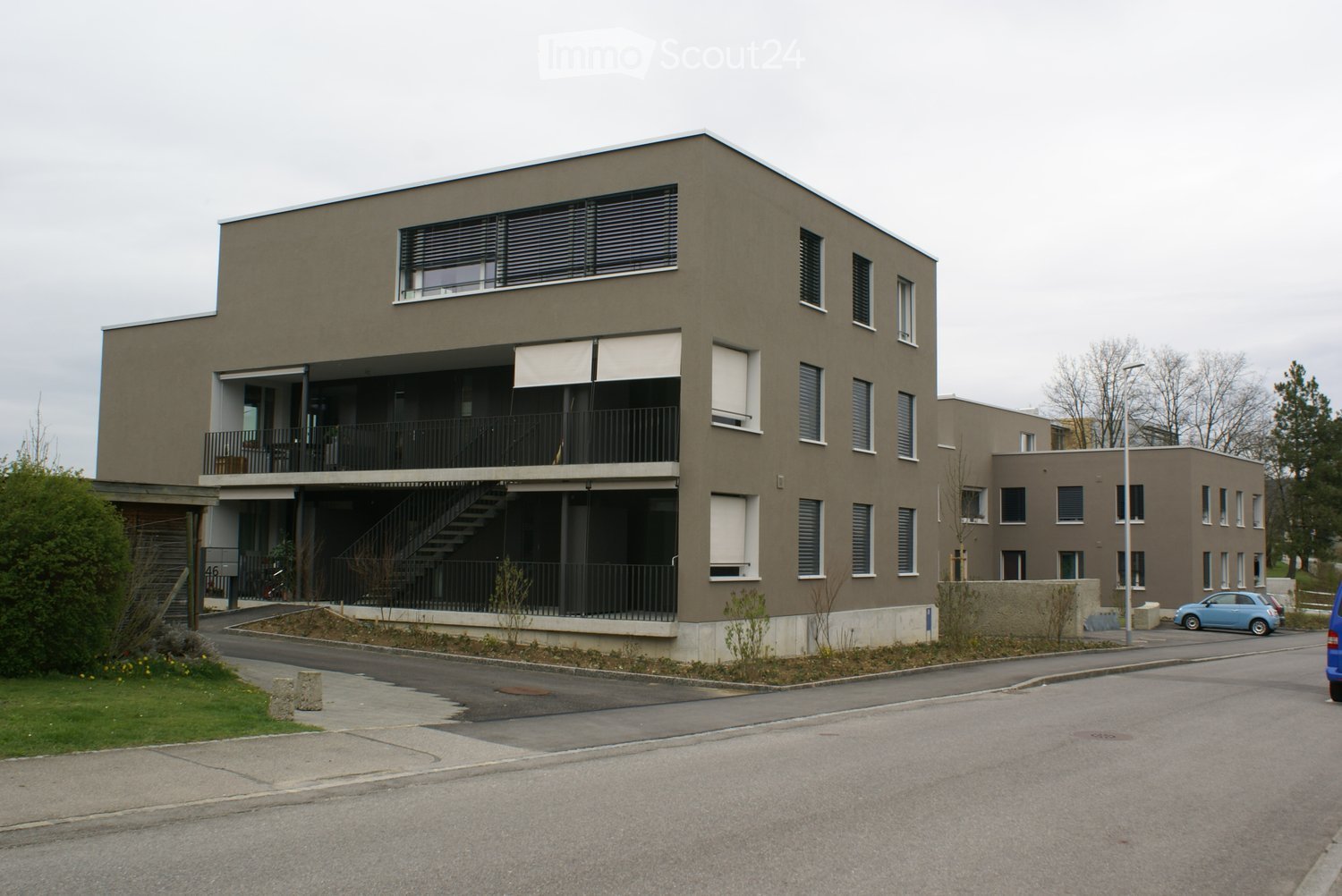 Modern two-story residential building with beige facade, large windows, balconies, and a staircase leading to the entrance.