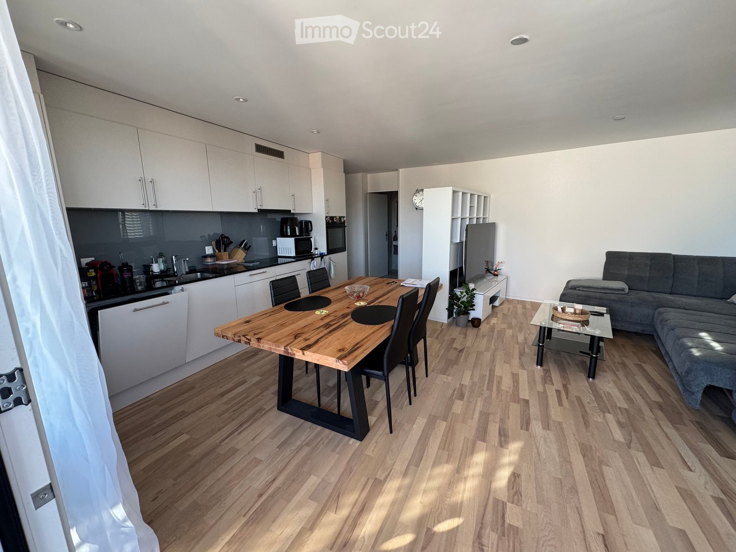 Modern open-plan living and dining area with light wooden flooring, white kitchen cabinets, a grey sofa, a glass coffee table, a wooden dining table with chairs, and a small plant near the TV stand.