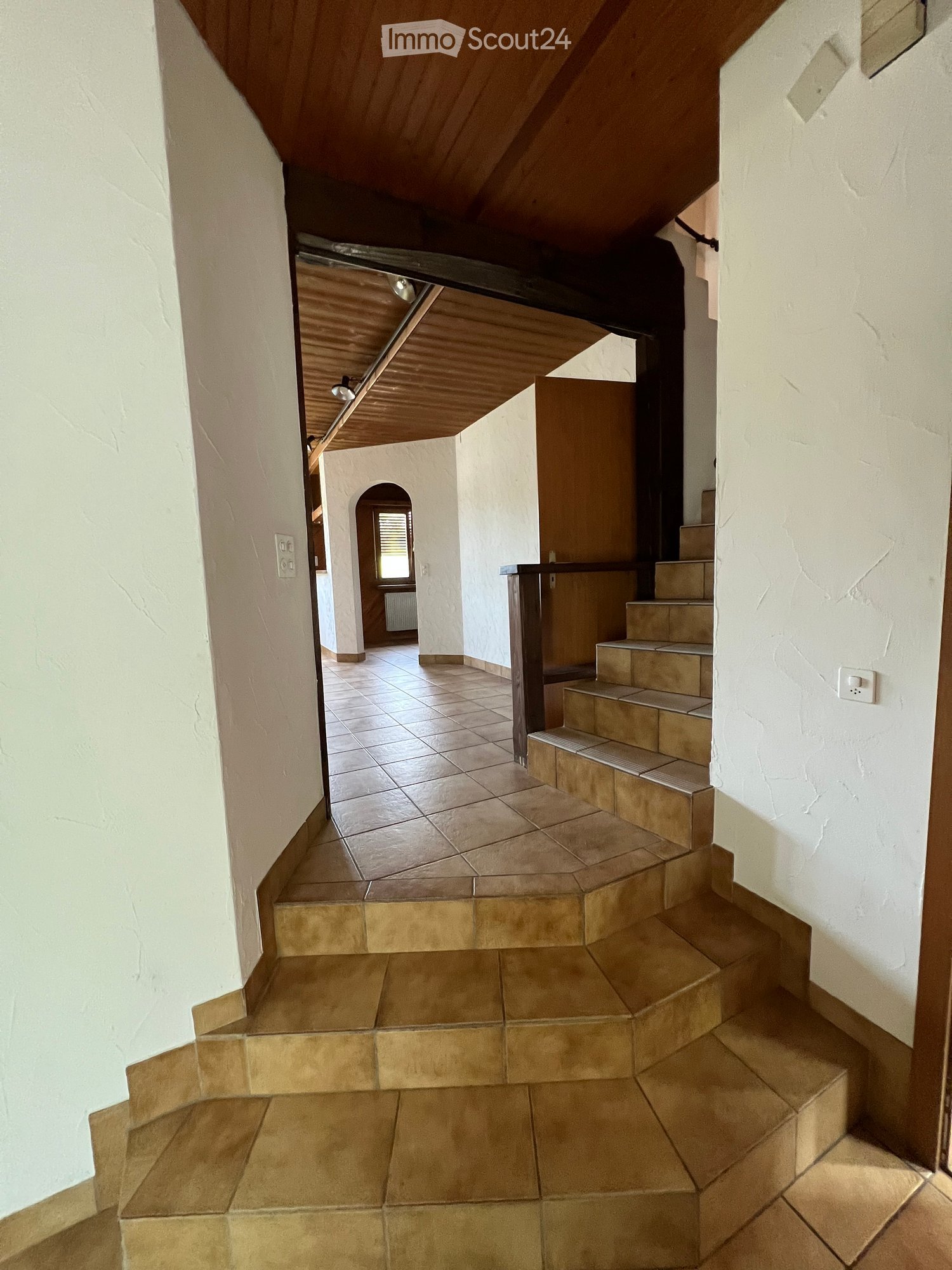 A hallway with tiled flooring and a wooden ceiling. There are white walls on both sides and a staircase leading to the upper floor. There is a power outlet on the left wall and a switch on the right wall. The hallway is empty.