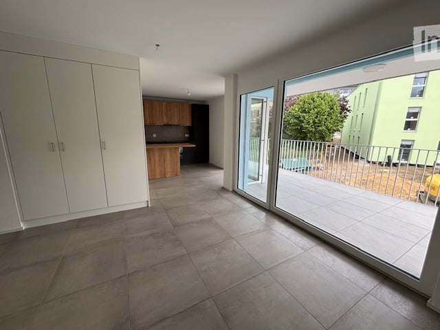 Empty room with tiled floor, large sliding glass doors leading to a balcony, white built-in cabinets, and a small kitchen area with wooden cabinets.