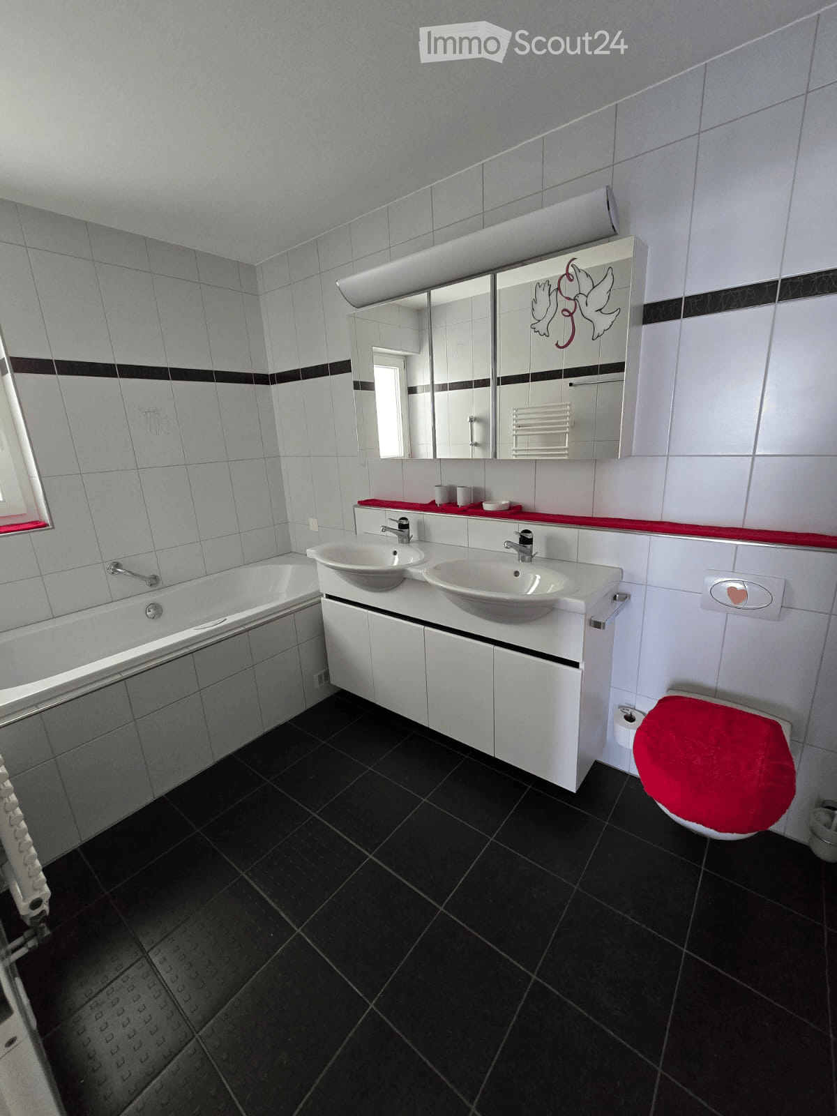 Modern bathroom with black tiled floor, white tiled walls, double sink vanity, bathtub, toilet with red seat cover, and a large mirror with decorative design.