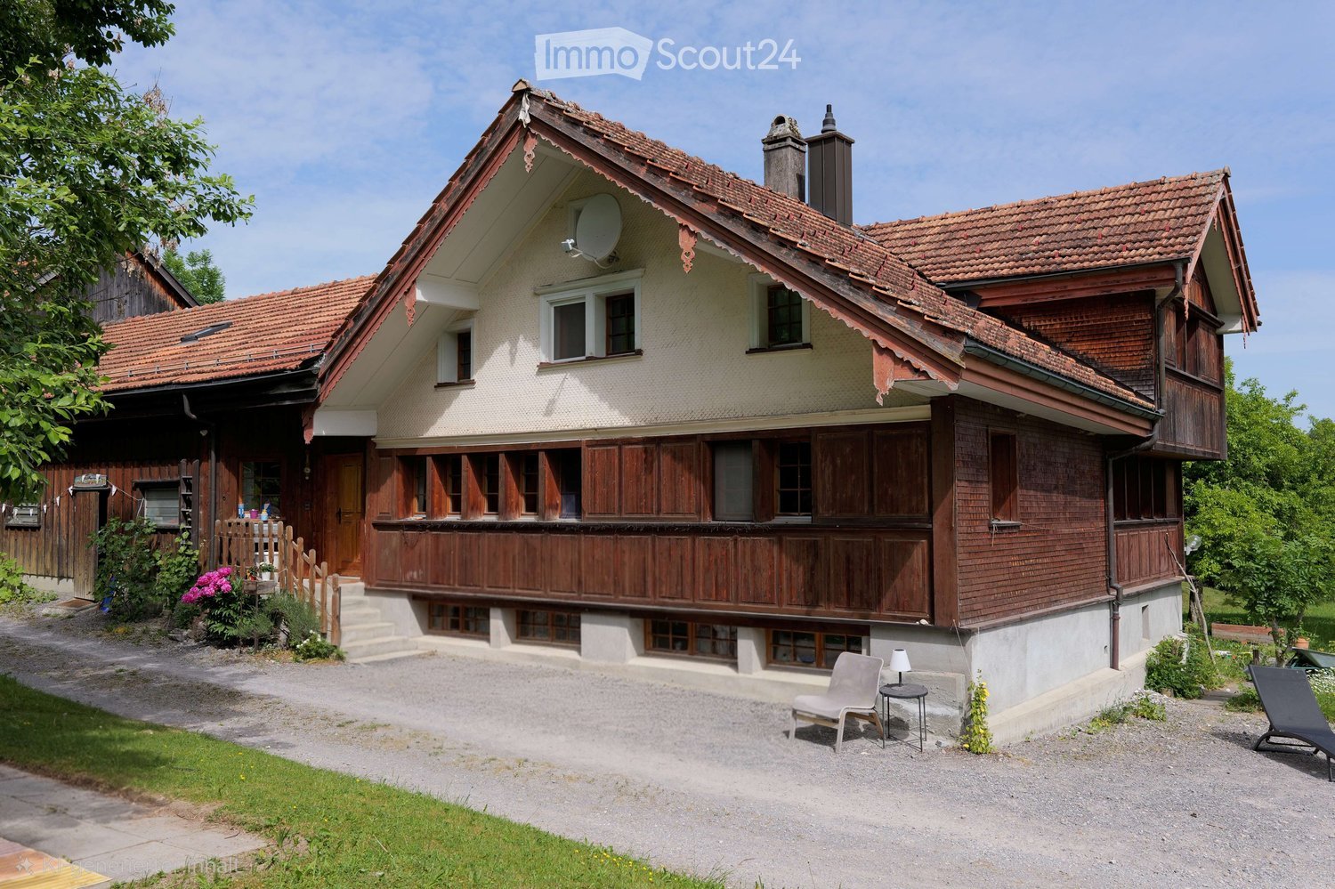 Two-story house with a red-tiled roof, wooden shutters, and a satellite dish. The house features a raised ground floor, a small porch with steps, and a gravel driveway with outdoor seating.