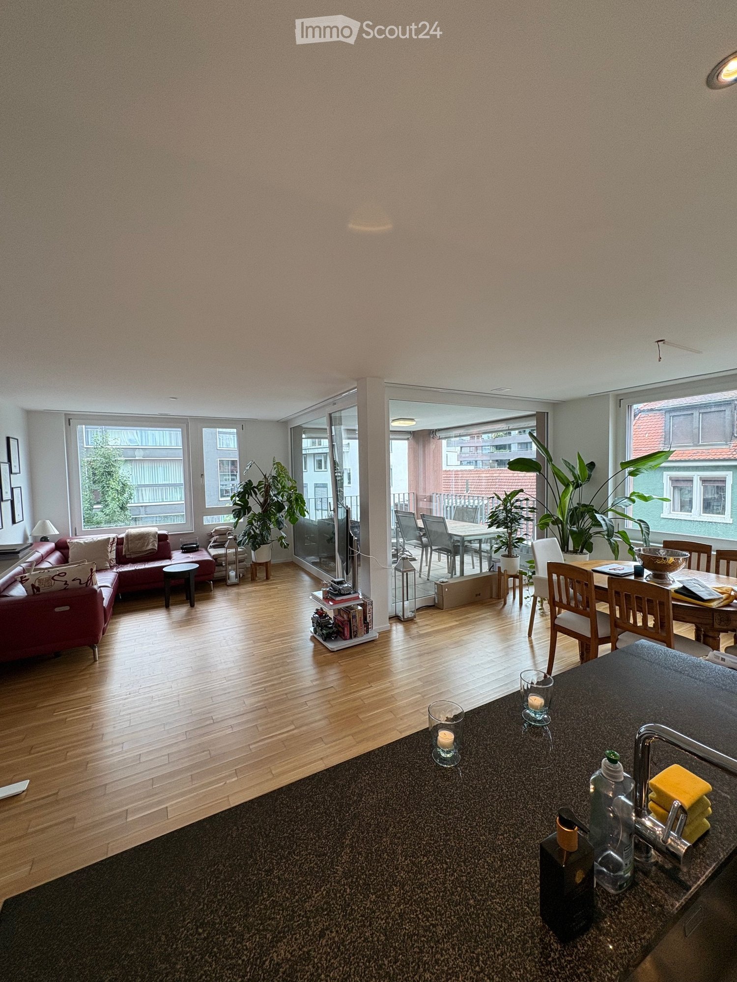 Open-plan living and dining area with wooden flooring, large windows, and a balcony. Features a red sofa, dining table with chairs, and a kitchen counter with a faucet and various items.