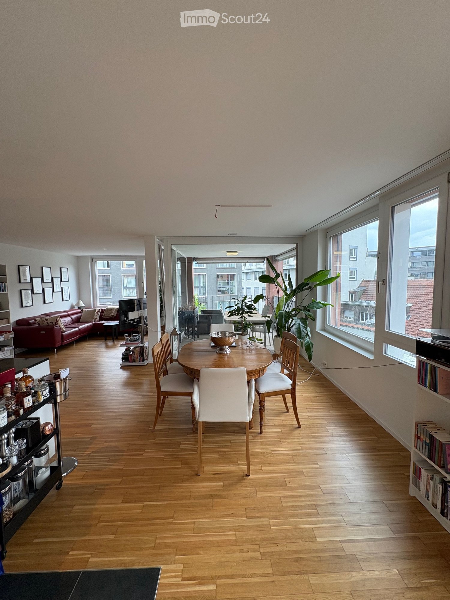 Open-plan living and dining area with wooden flooring, large windows, a red leather sofa, a round wooden dining table with chairs, and a bookshelf with books and decorative items.