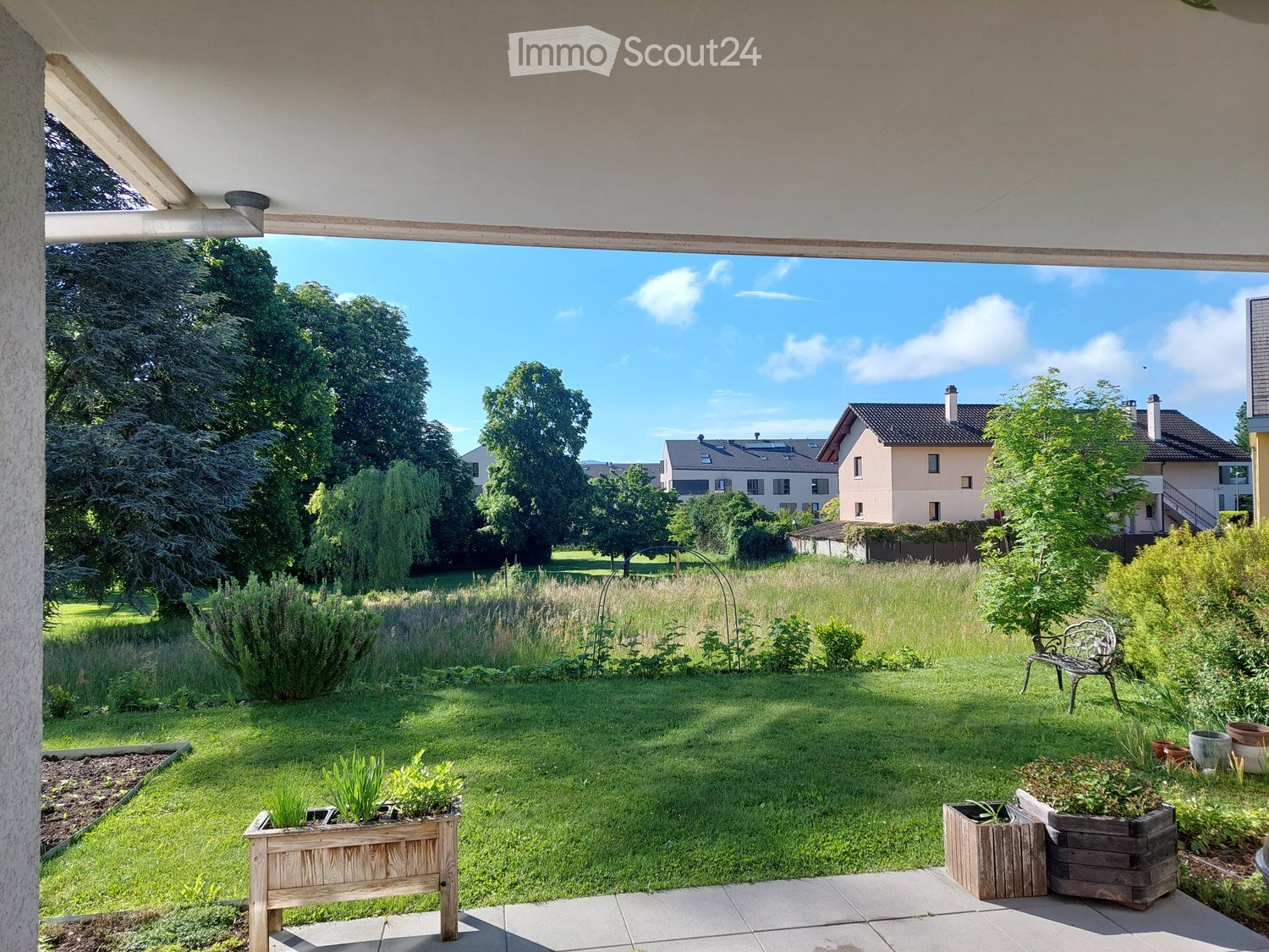 Well-maintained garden with a grassy lawn, wooden planters, and a metal bench, overlooking a residential area with trees and houses.