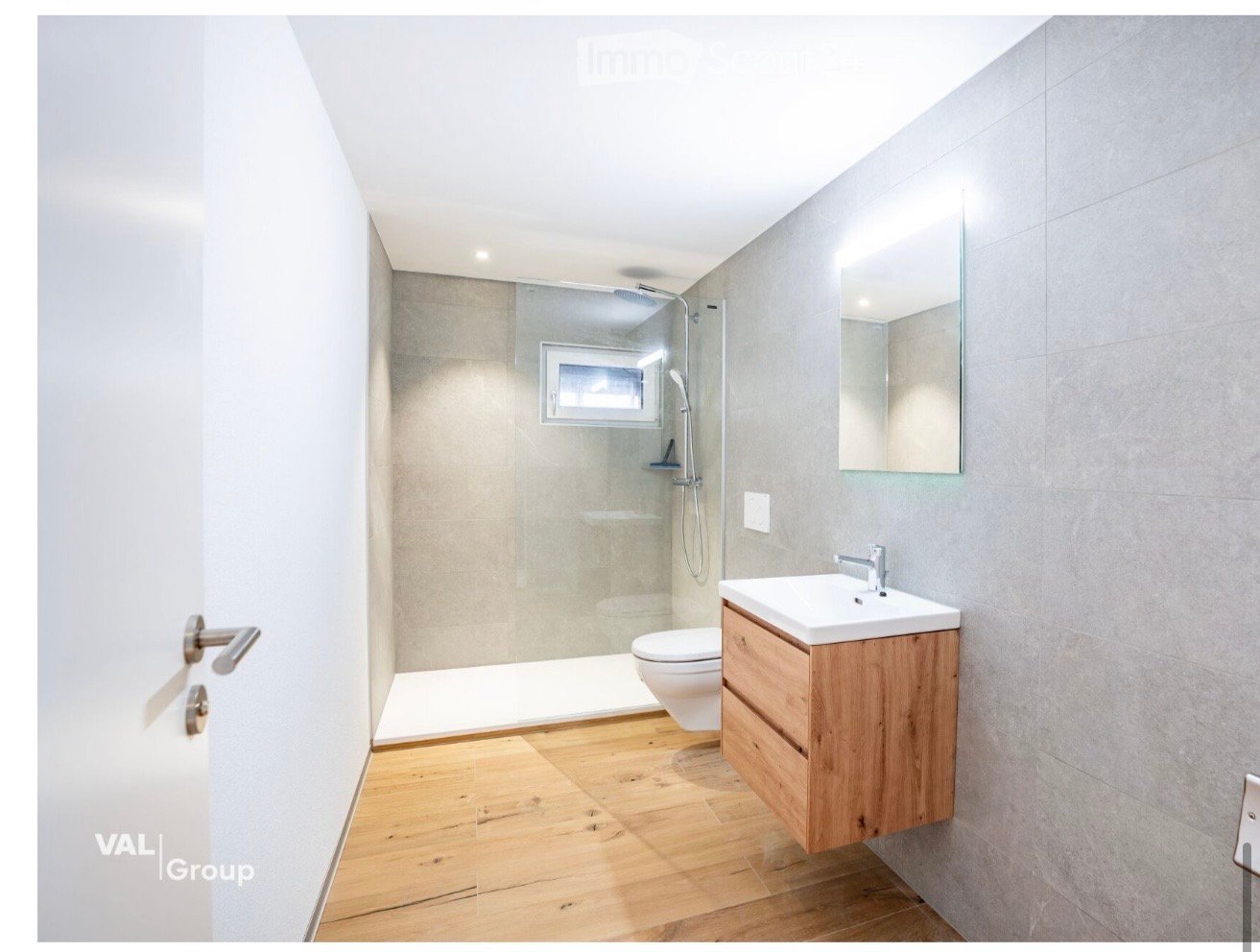 Modern bathroom with wooden flooring, a white sink with wooden cabinet, a toilet, a mirror, and a glass-enclosed shower area.