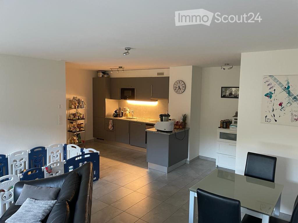 Open-plan living and dining area with a modern kitchen. Features include a grey sofa, dining table with chairs, kitchen cabinets, microwave, clock, and a small shelf with items.