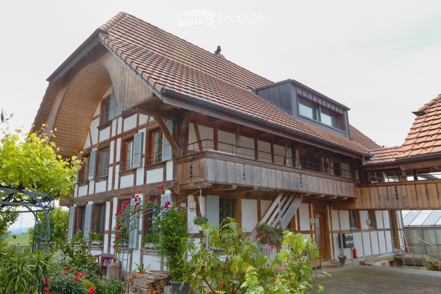 Traditional two-story house with wooden beams and tiled roof, featuring a balcony and surrounding garden with plants and flowers.