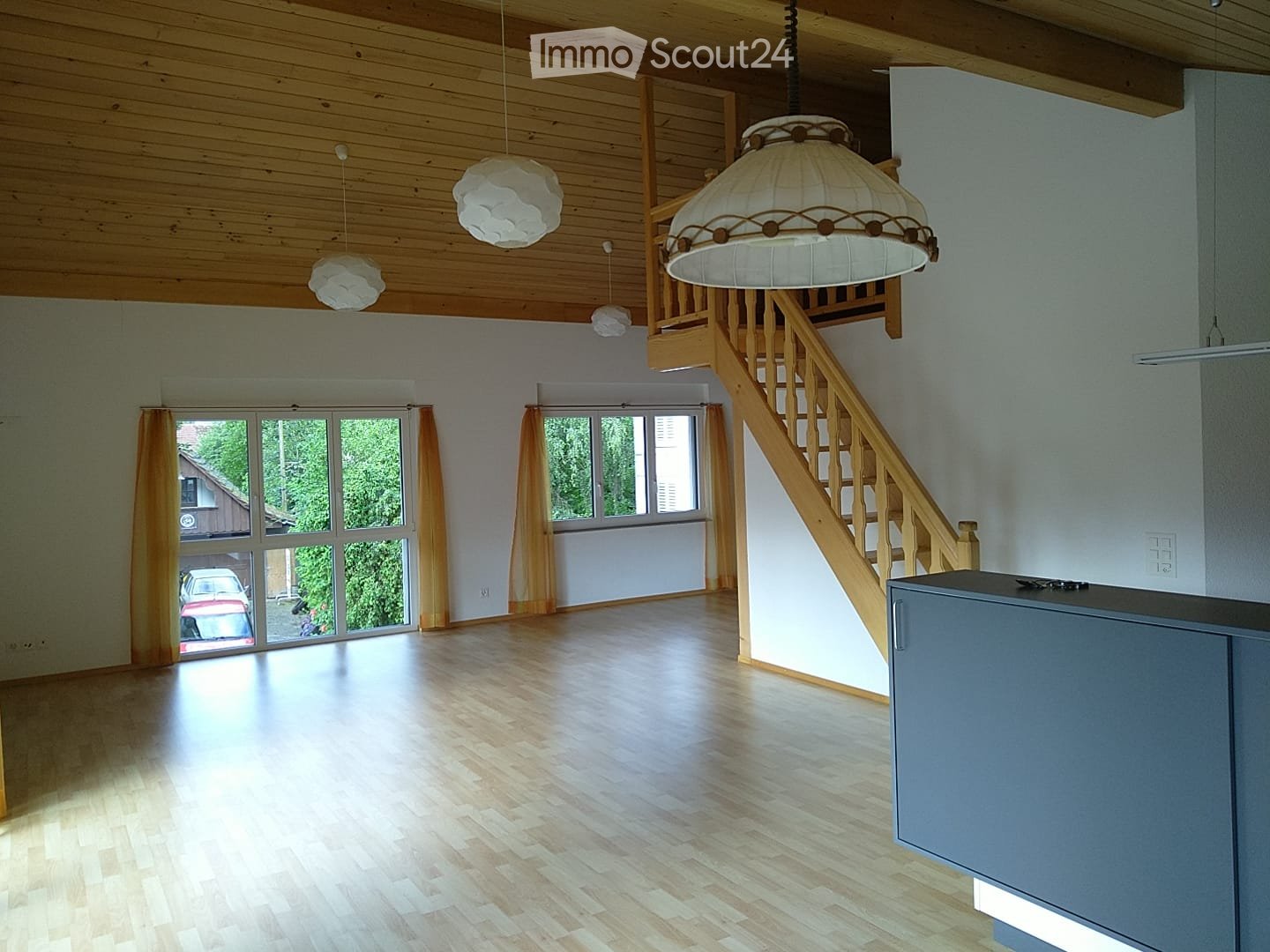 Bright, empty room with wooden flooring, large windows with yellow curtains, wooden staircase leading to an upper level, and several hanging lamps.