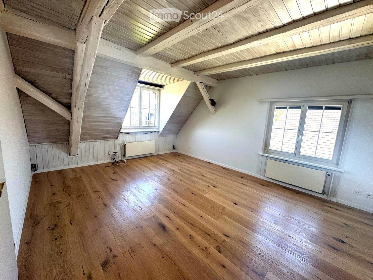 Empty attic room with wooden floors, white walls, exposed wooden beams, and two windows with blinds.
