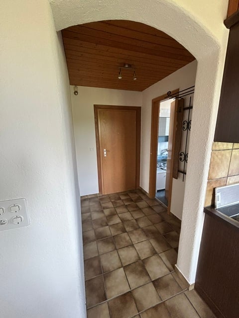 A narrow hallway with tiled flooring, wooden ceiling, and a closed wooden door on one side. An open doorway leads to another room with visible furniture.