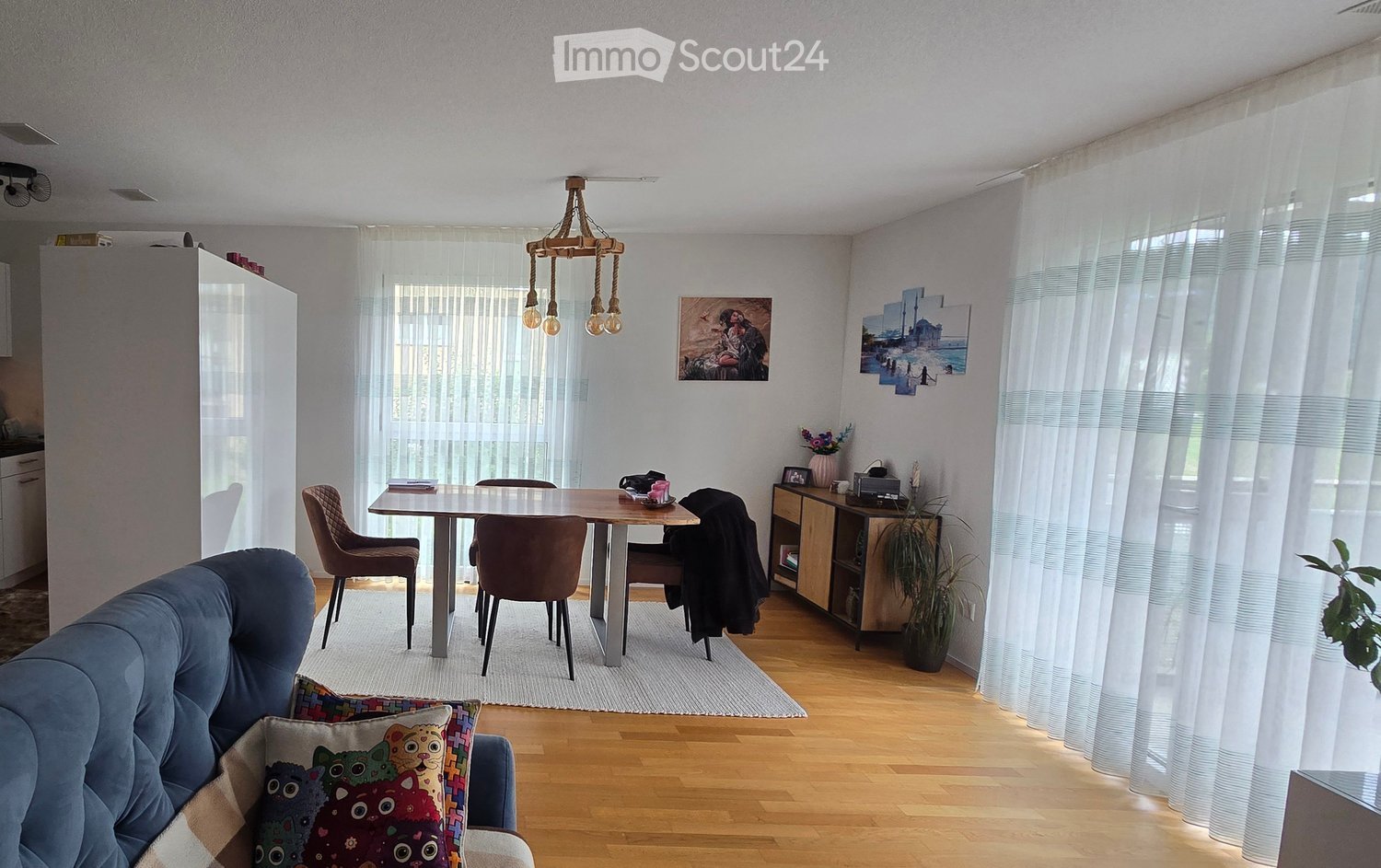 A modern living and dining area with a blue sofa, colorful pillows, a wooden dining table with chairs, a chandelier, and large windows with white curtains.