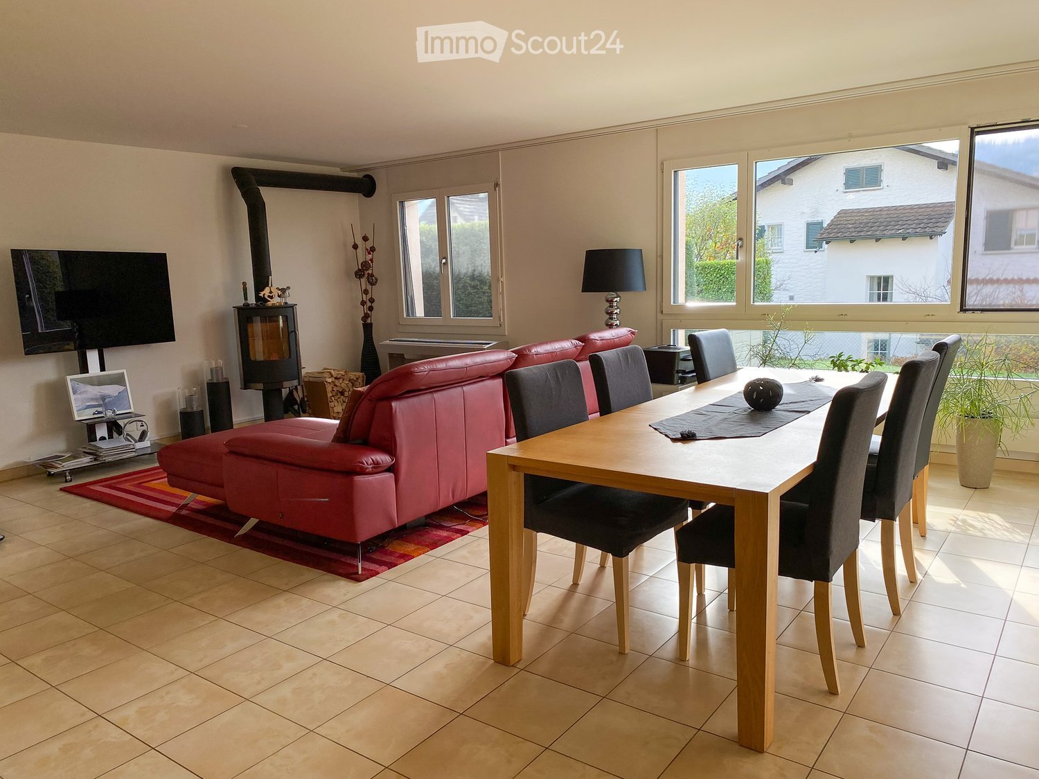 A modern living and dining area with a red leather sofa, a wooden dining table with chairs, a fireplace, a lamp, and a TV screen. The room has large windows and a view of the outside.