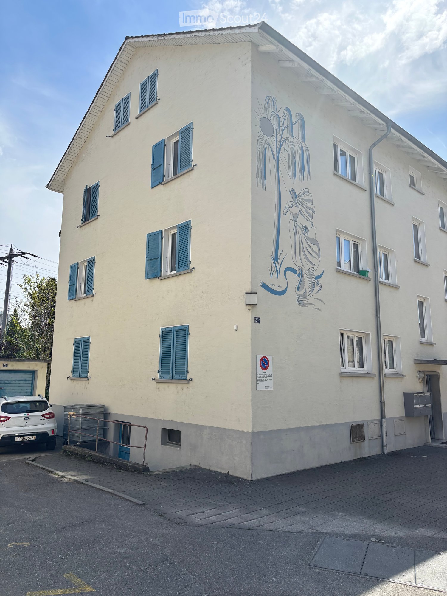 Two-story building with light yellow walls, blue shutters, and a mural of a mermaid on one side. Features a garage, a small staircase, and a parked car.
