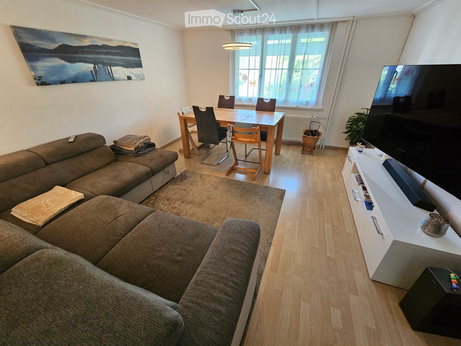 A modern living and dining area with a sectional sofa, coffee table, dining table with chairs, TV, and a plant. The space features wooden flooring and large windows with curtains.