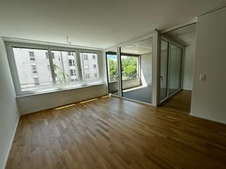 Completely empty room with wooden flooring, large windows, and a sliding glass door leading to a balcony.