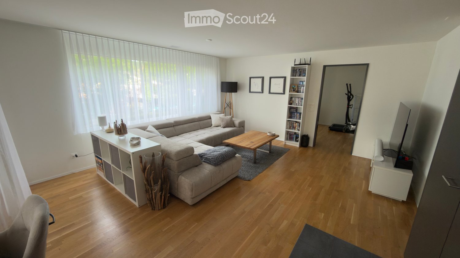 Modern living room with light wooden flooring, large windows with sheer curtains, grey sectional sofa, coffee table, bookshelf, floor lamp, and exercise bike in the background.