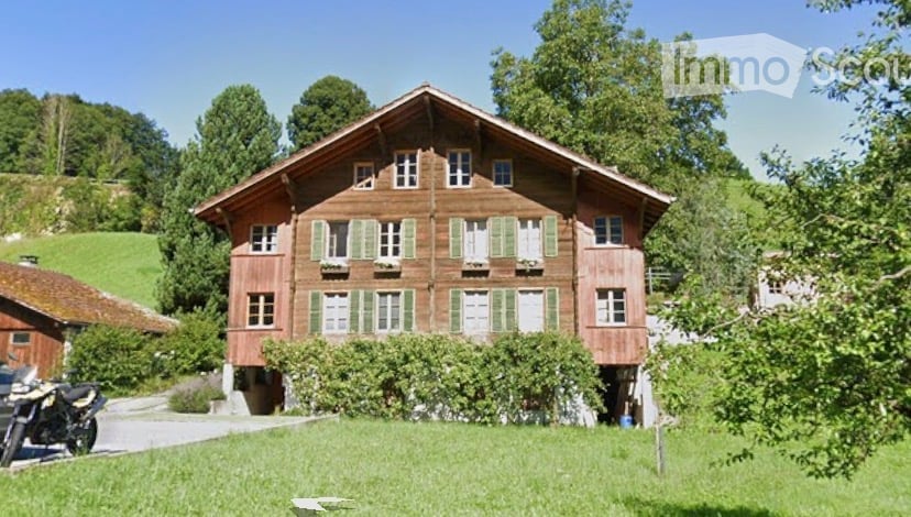 Traditional wooden house with green shutters and multiple windows, surrounded by trees and greenery.