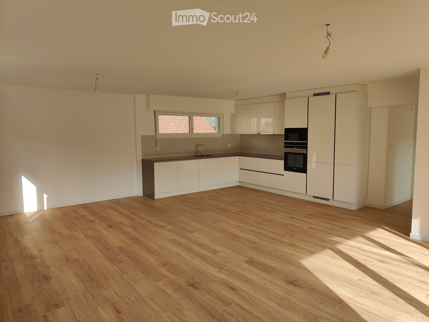 Empty kitchen with white cabinets, wooden flooring, and natural light from windows. No furniture or appliances are present.