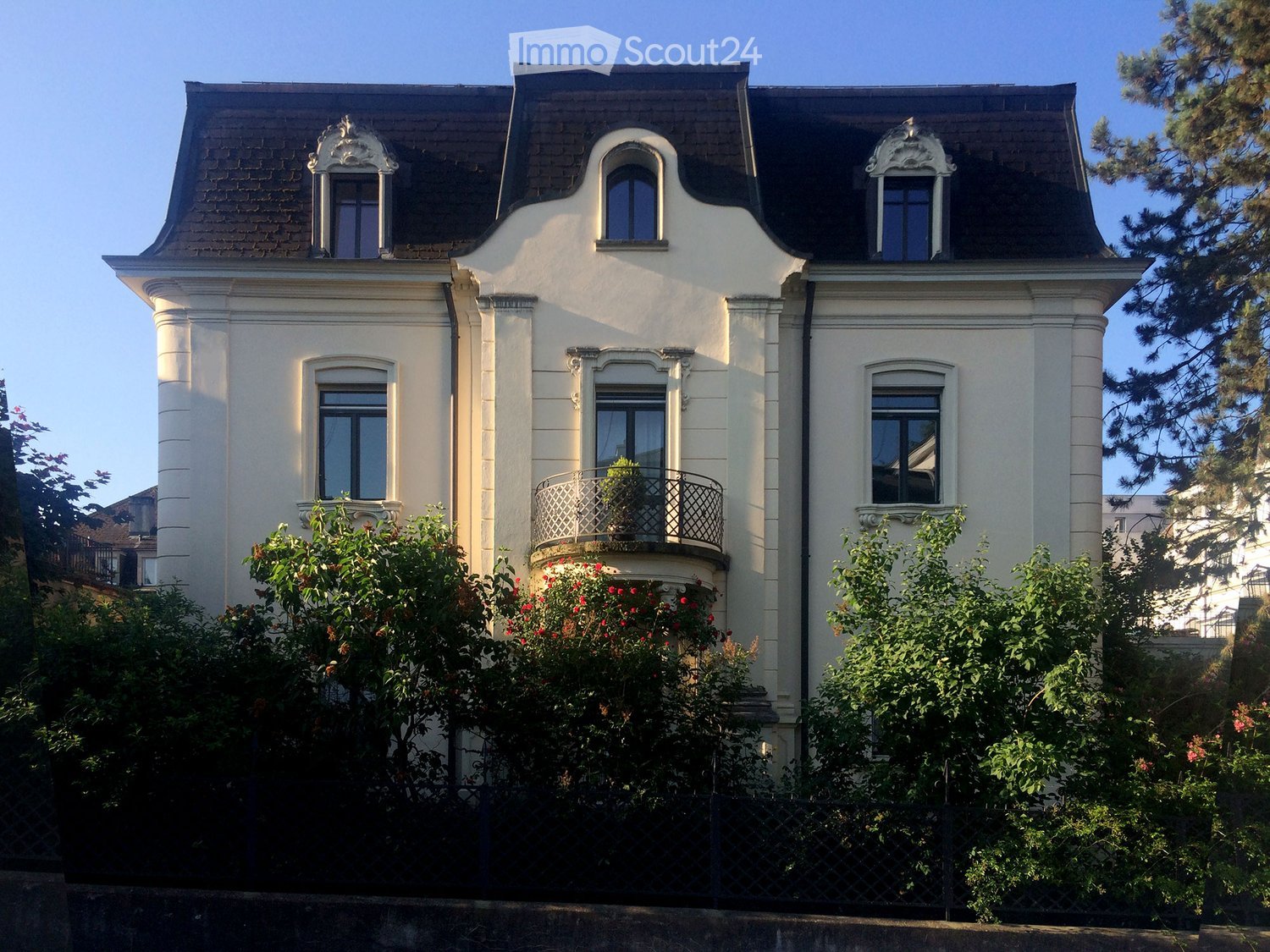 A three-story white house with ornate architectural details, including decorative window surrounds and a small balcony with a potted plant. The house features multiple windows and is surrounded by lush greenery and flowering bushes.