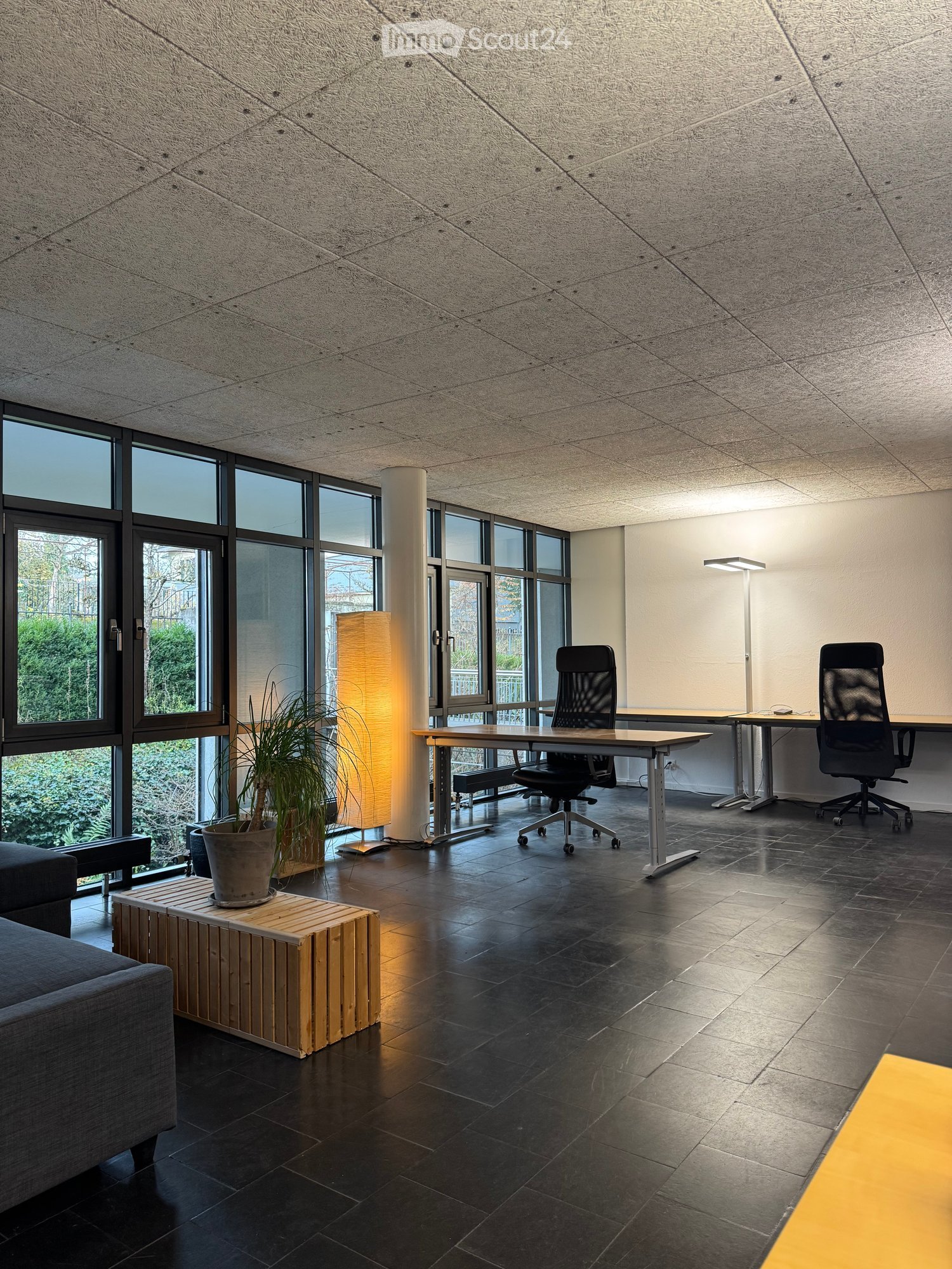 Modern office space with dark tiled flooring, large windows, a sofa, a small wooden table with a potted plant, two desks with chairs, and a floor lamp.