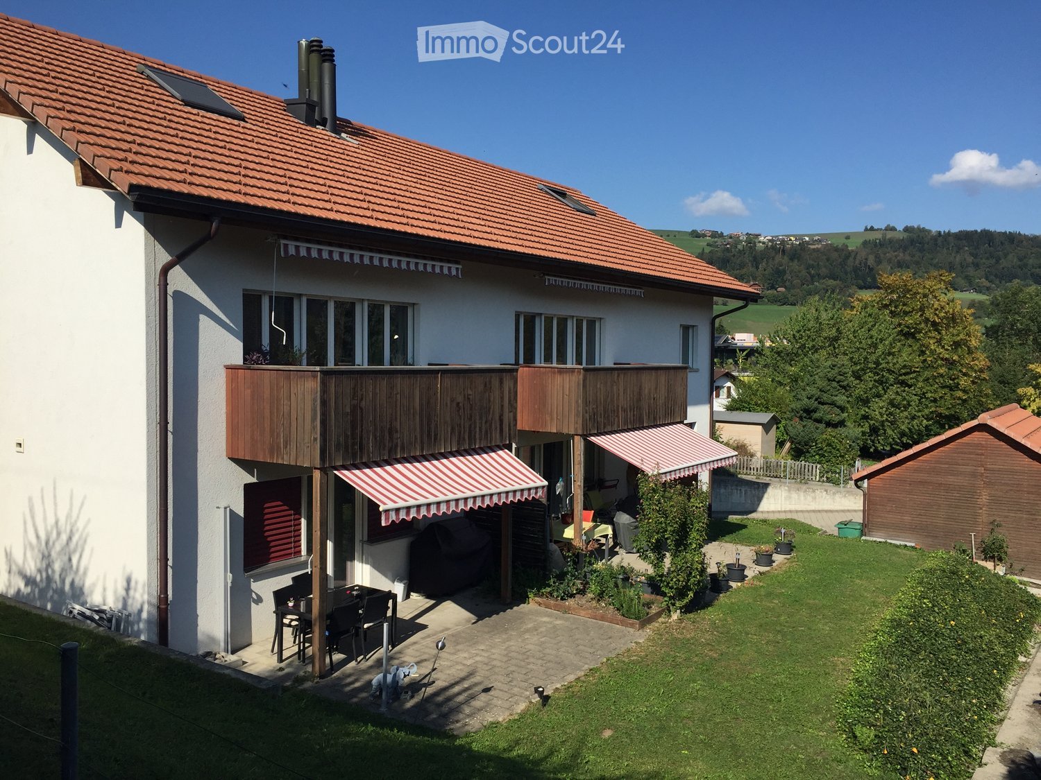 Two-story house with red-tiled roof, white walls, multiple windows, and a wooden balcony. The house features striped awnings, a patio with outdoor furniture, and a well-maintained garden with lawn and potted plants.