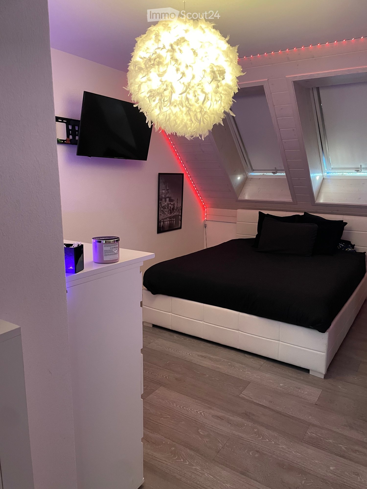 Modern bedroom with a white platform bed, black bedding, a hanging feather lamp, a nightstand with a lamp, and a TV mounted on the wall.