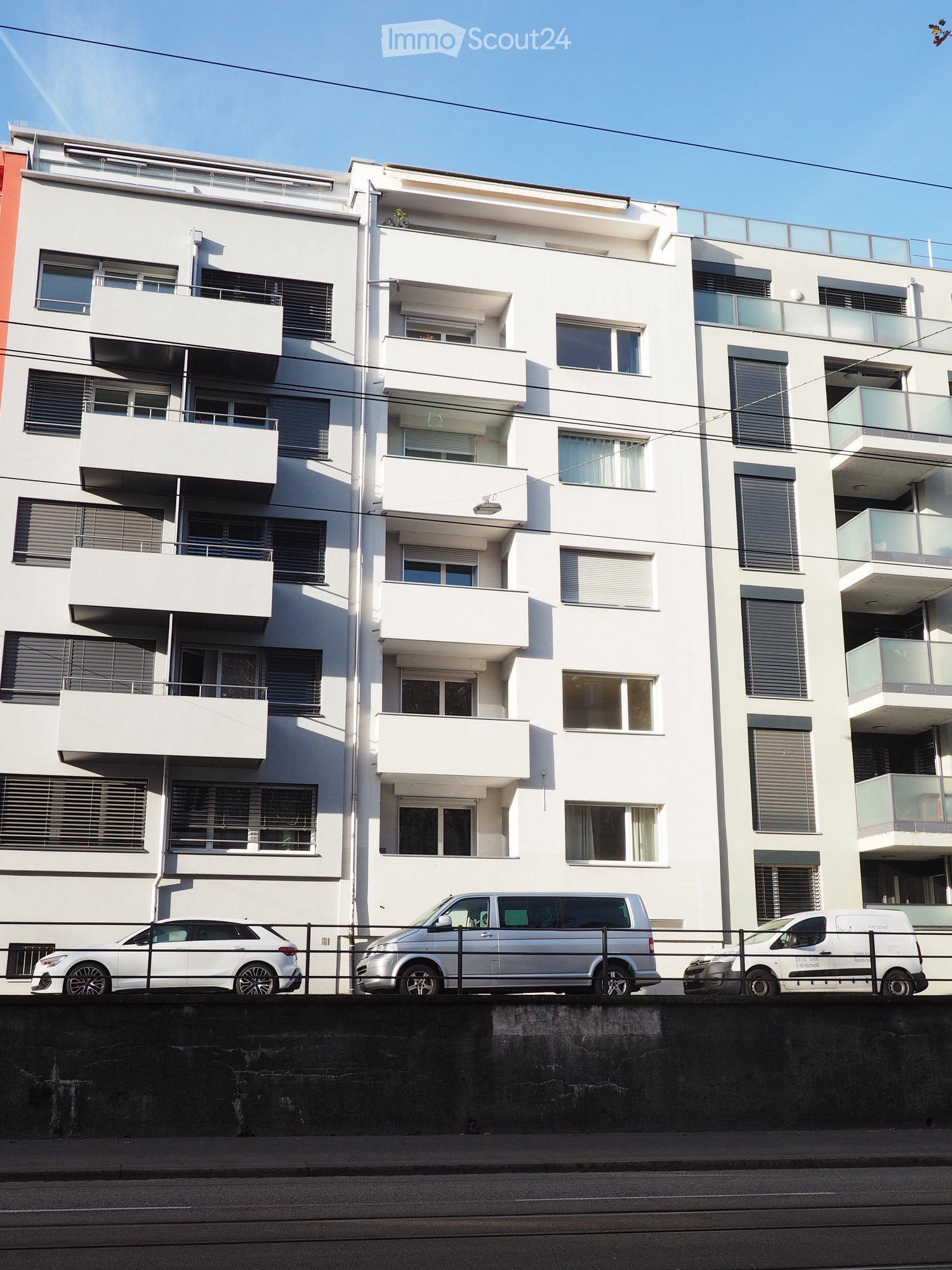 Modern multi-story residential building with white facade, multiple balconies, and large windows. Several cars are parked in front.