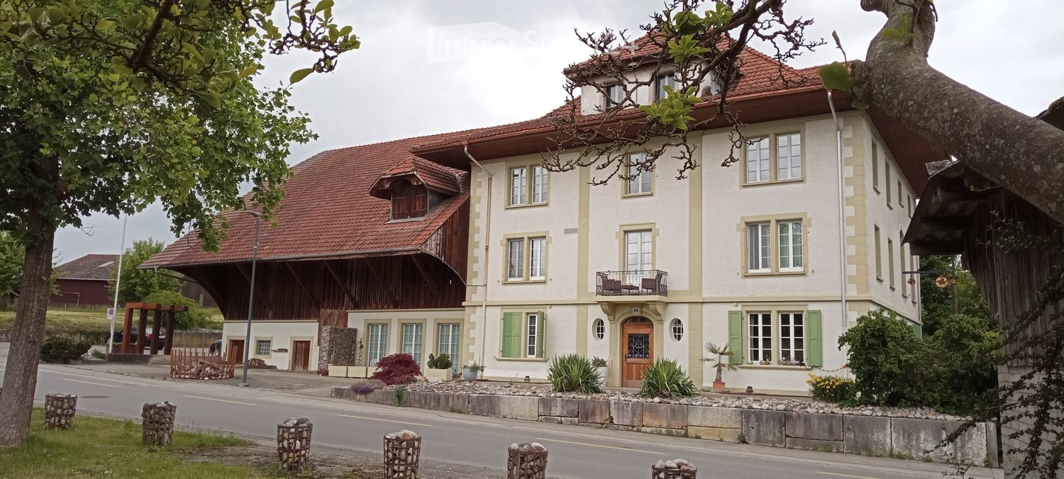 A two-story building with a red-tiled roof, white walls, green shutters, and a wooden balcony. The building has multiple windows and a central entrance with a small porch.
