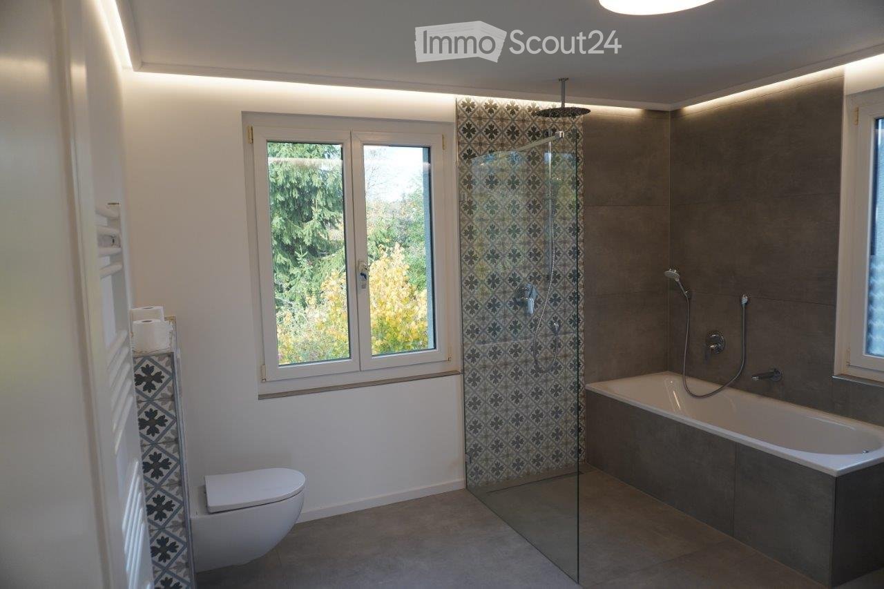 Modern bathroom with a bathtub, shower, toilet, and towel rack. Features large windows with a view of greenery, patterned tiles, and built-in lighting.