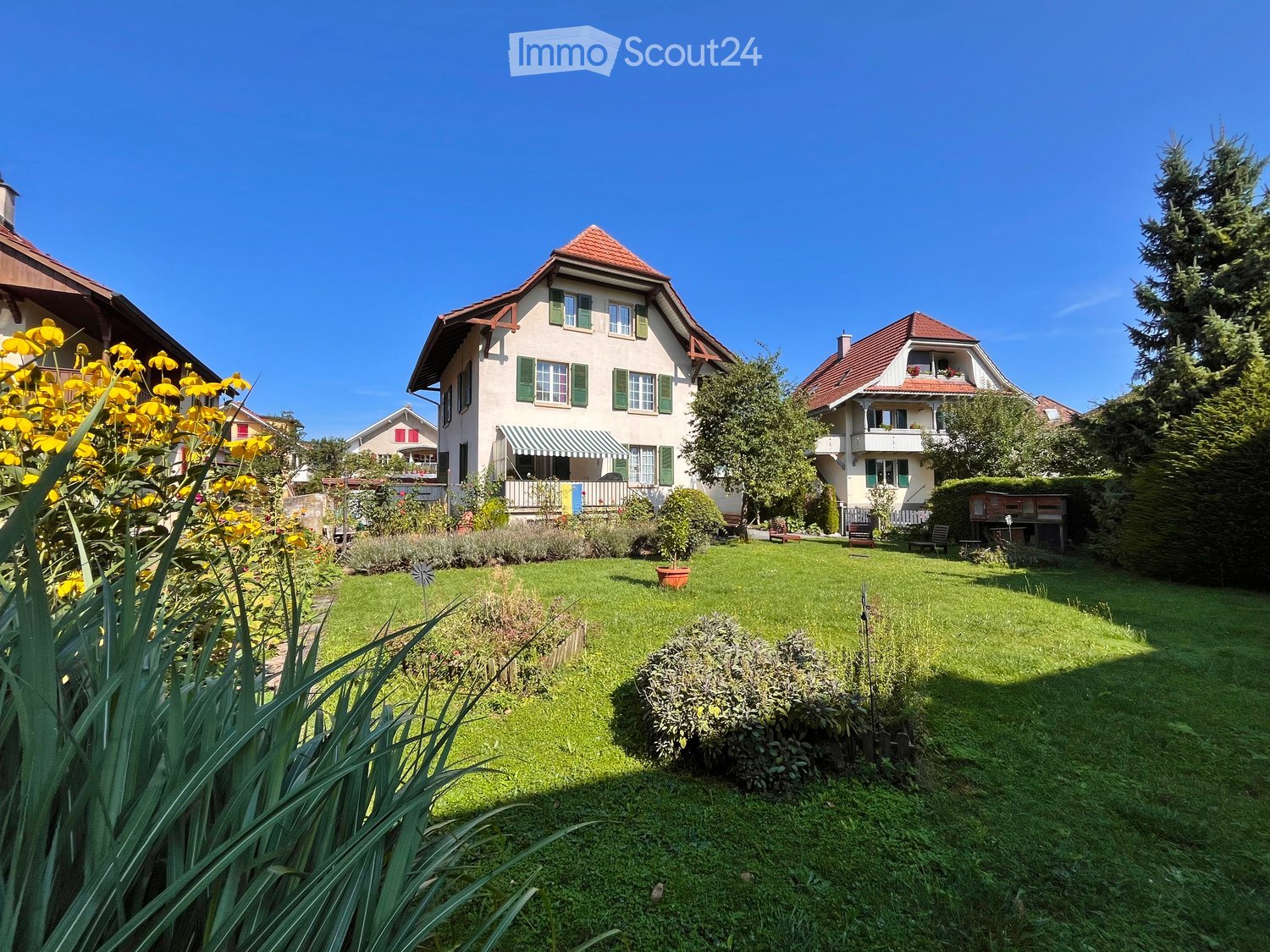 Well-maintained garden with green lawn, flower beds, shrubs, and surrounding houses with red-tiled roofs and green shutters.