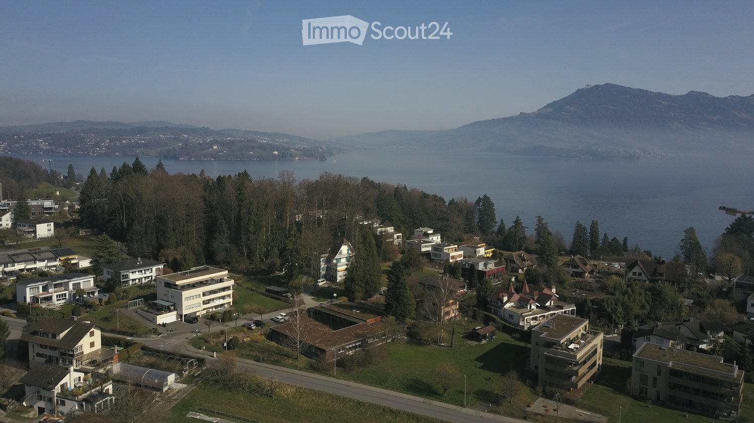 Aerial view of a residential area with multiple buildings, surrounded by trees and a lake in the background.