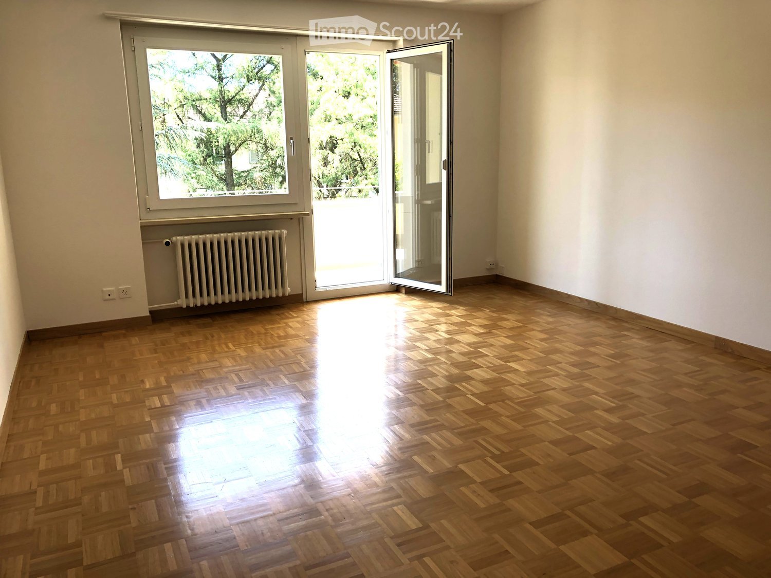 Empty room with wooden parquet flooring, white walls, large window with open sliding doors leading to a balcony, radiator, and visible electrical outlets.