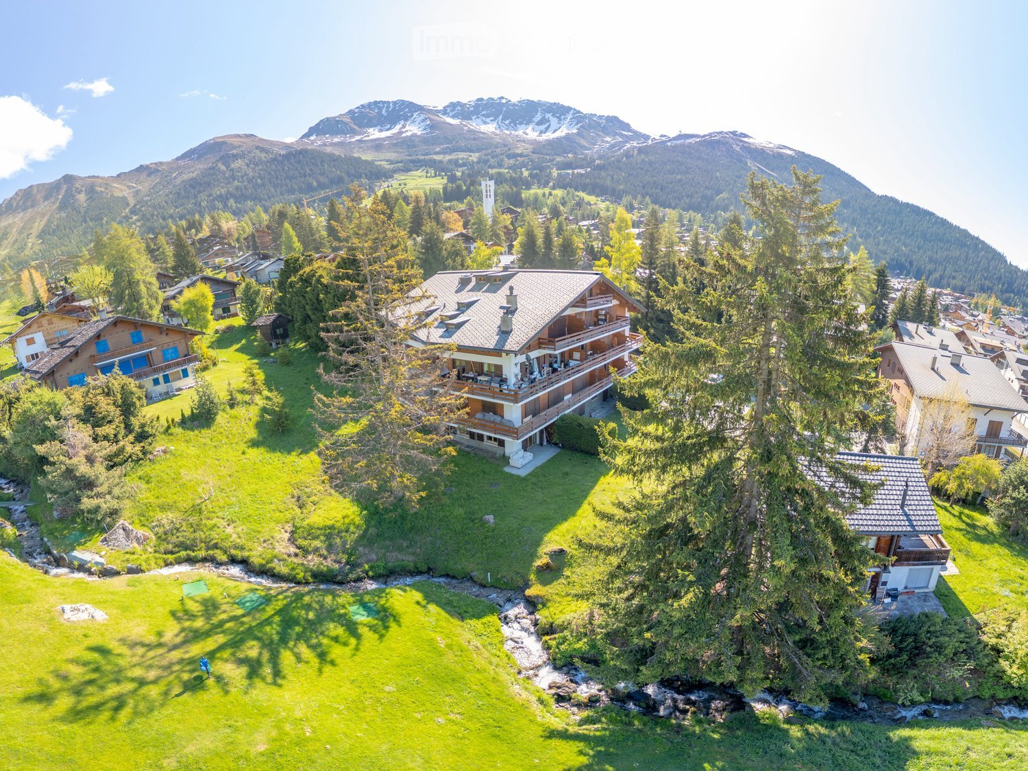 A large, multi-story house with a wooden exterior and multiple balconies, surrounded by lush green lawns, trees, and a small stream, with snow-capped mountains in the background.