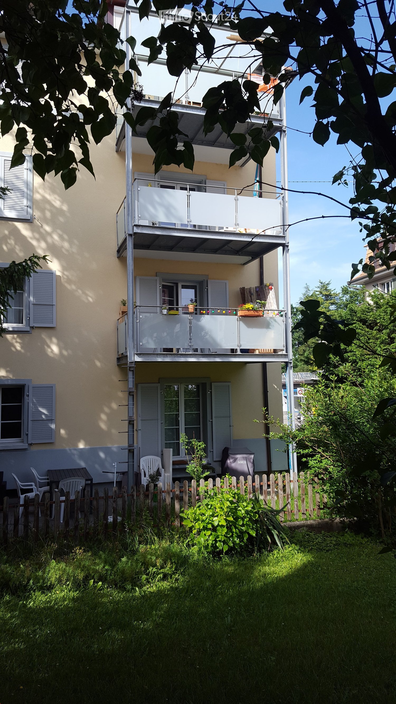 Balcony with glass railing, outdoor furniture, and potted plants, surrounded by greenery and a wooden fence.