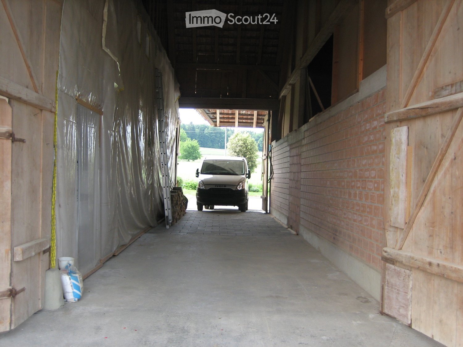 A wide, open corridor with wooden walls and a concrete floor, leading to an outdoor area with a parked van visible through the doorway.