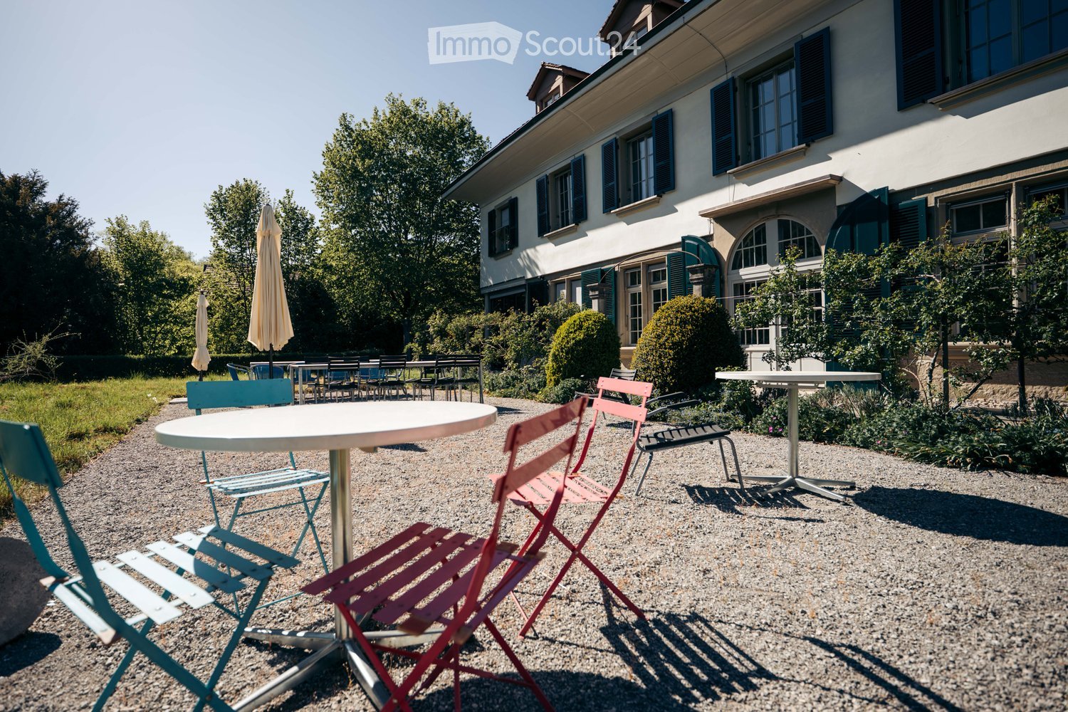 Outdoor seating area with tables and chairs, surrounded by greenery and a building with shutters in the background.