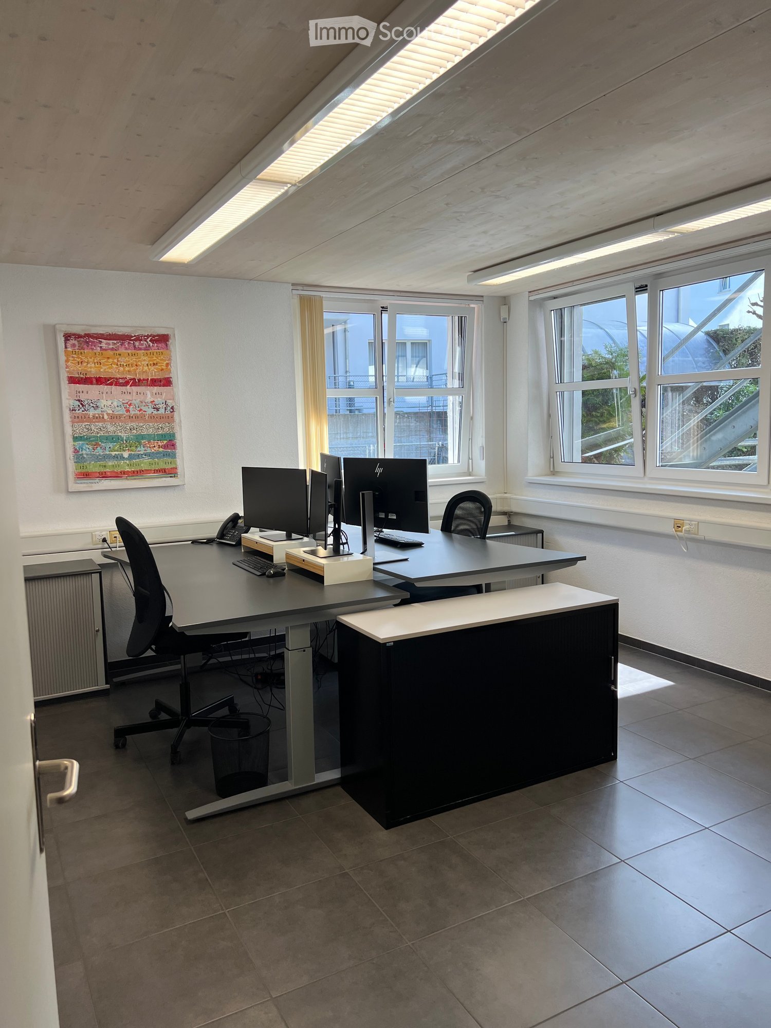 Office space with desks, chairs, computers, and a reception desk, large windows, and a slanted ceiling with skylights.