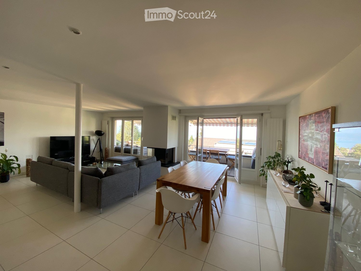 Open-plan living and dining area with a grey sofa, wooden dining table, white chairs, television, floor lamp, and plants. Large windows and sliding glass doors lead to a balcony.
