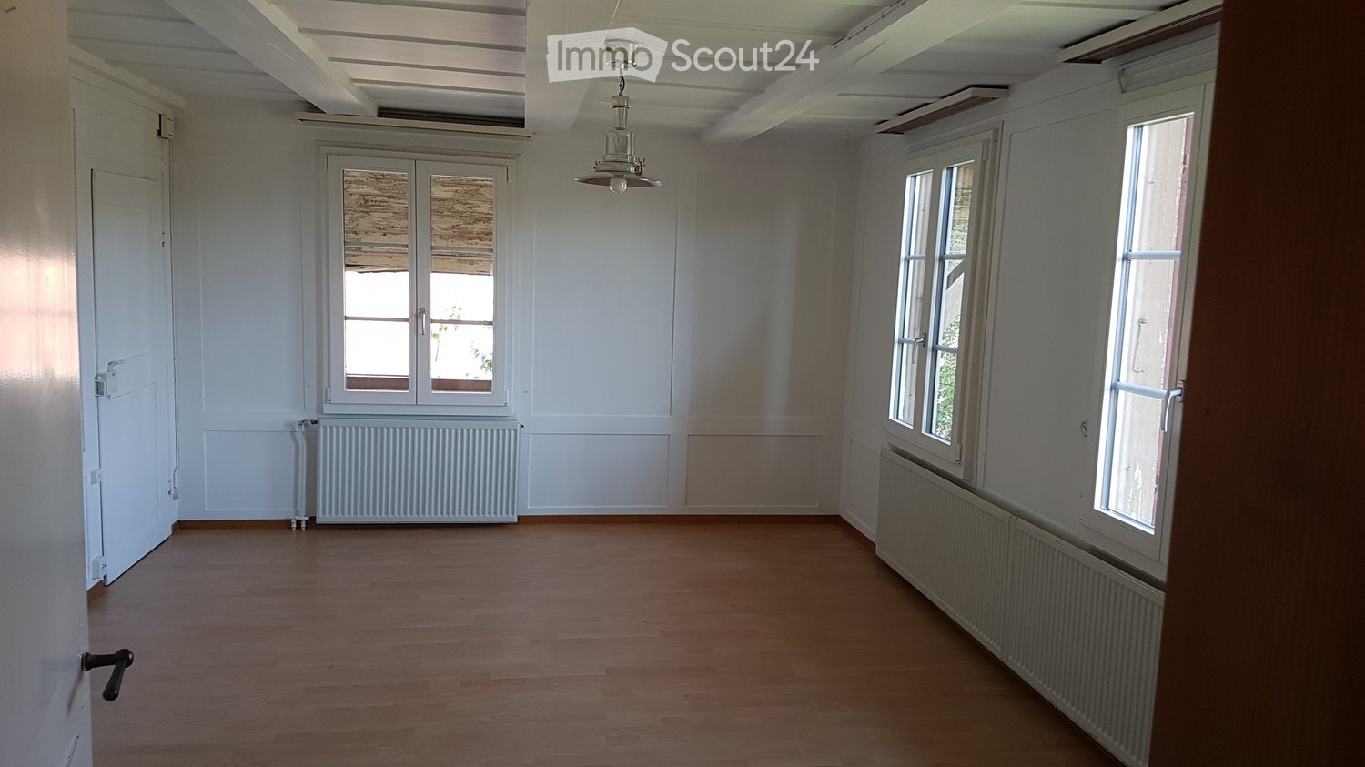 Empty room with wooden flooring, white walls, large windows, radiators, and a hanging light fixture.