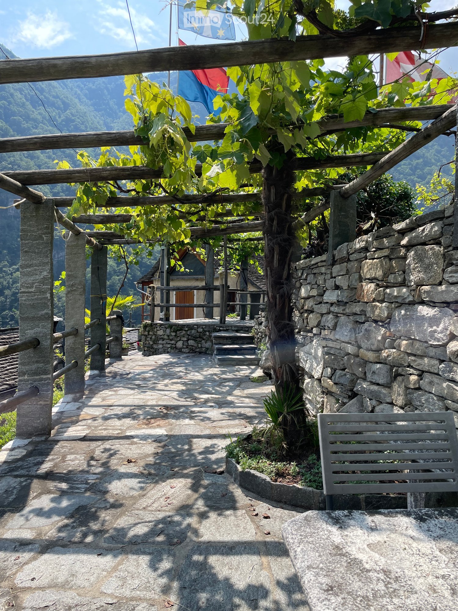 A stone-paved outdoor area with a wooden pergola, stone walls, a bench, and a view of a mountainous landscape.